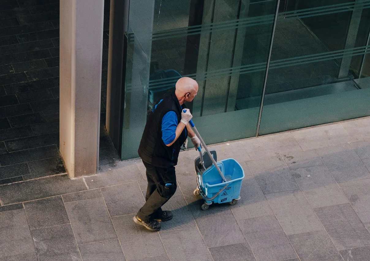 Cleaner wheeling a mop bucket along the paving outside a glass office building